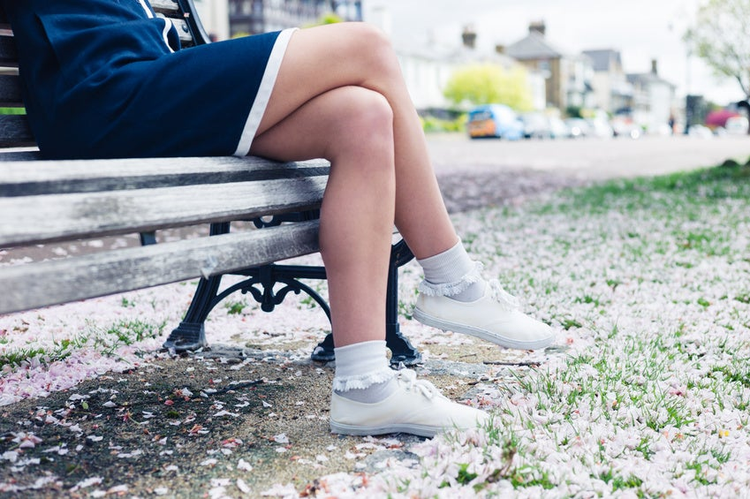The legs and feet of a young woman relaxing on a park bench with cherry blossom on the ground