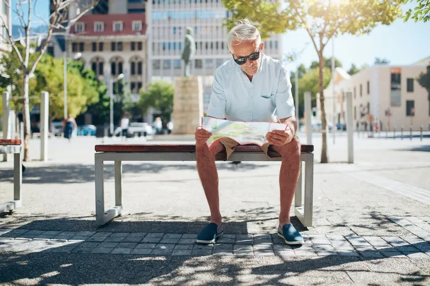 Senior male tourist sitting on a bench and looking for a place on the map. Mature man sitting outdoors in the city and reading a map on a summer day.