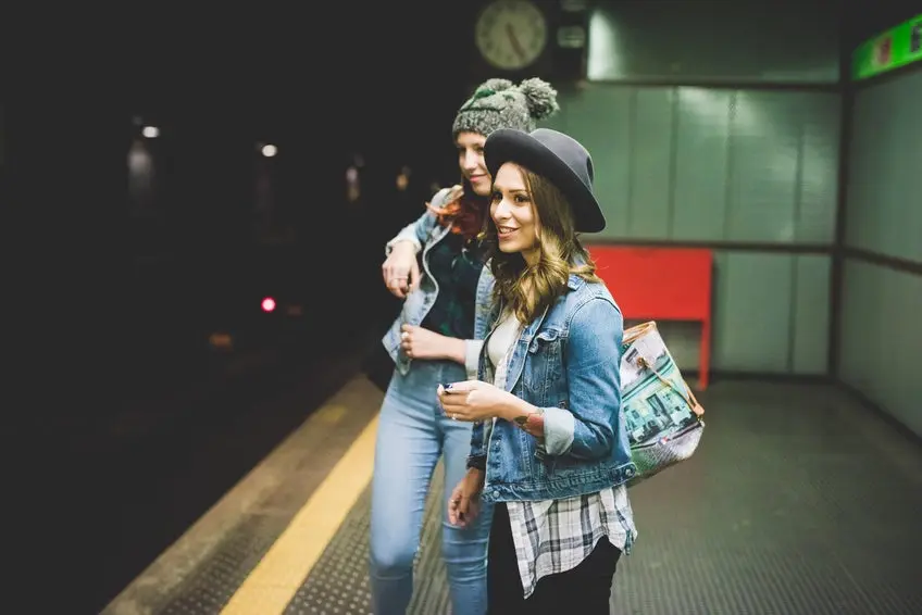 Knee figure of two young handsome caucasian blonde and redhead straight hair women waiting in the underground for tube, one using smartphone - transport, technology concept