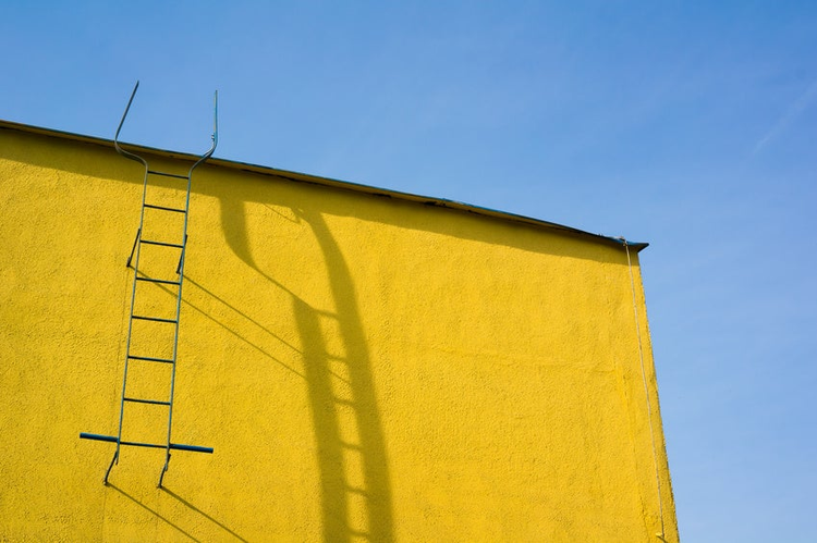 Top of yellow wall. Ladder to the roof and its shadow. Blue sky.