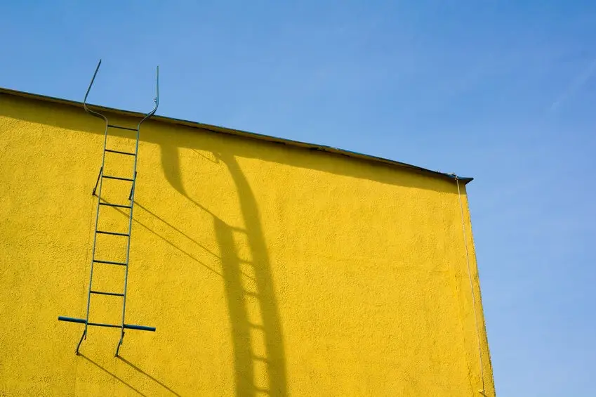 Top of yellow wall. Ladder to the roof and its shadow. Blue sky.