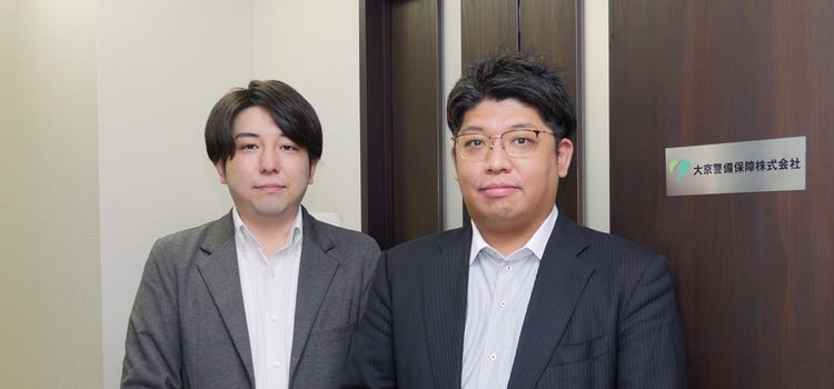 白い壁の前に立つスーツを着た男性 中程度の精度で自動的に生成された説明