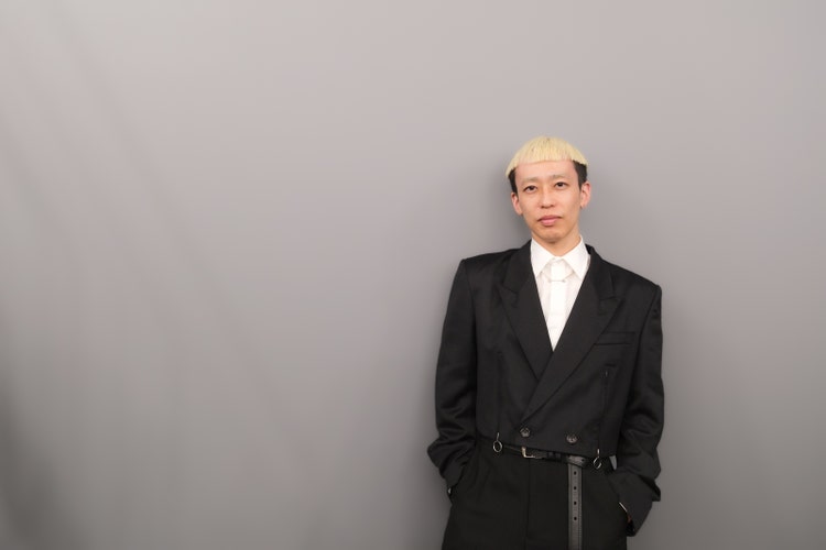 白い壁の前に立つスーツを着た男性 中程度の精度で自動的に生成された説明