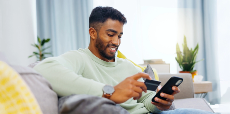 Man sitting on a couch, smiling while using a smartphone and holding a credit card.