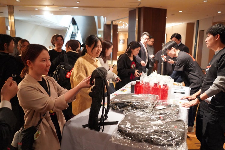 Corporate Social Responsibility（CSR）活動の一環で、チャリティ販売を実施している写真です。ウォーターボトルや、リュックなど、アドビのロゴ入りのグッズを手にした社員たちが、どれにしようか迷ってます。