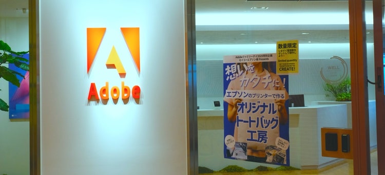 Adobeロゴが掲示されたオフィス入口と、オリジナルトートバッグ制作を案内する展示パネル